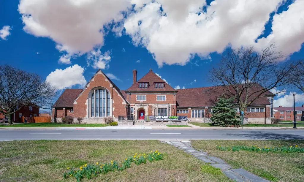 Wide-angle view of the historic Parkman Library Branch in Detroit, featuring Tudor Revival architecture, red brick façade, and arched windows under a clear blue sky.