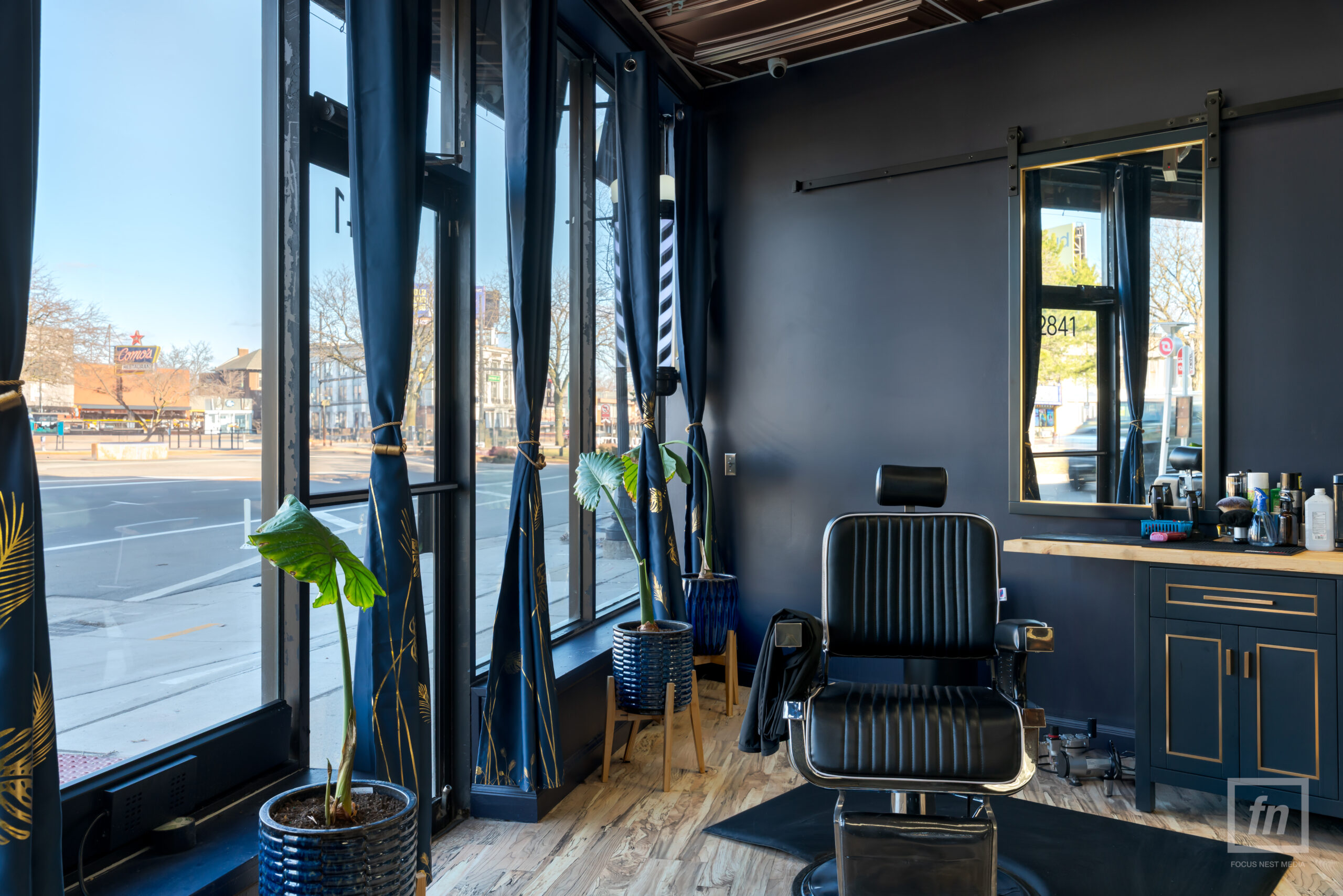 Barber station at The Braxton Grooming Lounge in Ferndale with black barber chair, brass-trimmed mirror, blue cabinetry, plants, and street-facing windows.