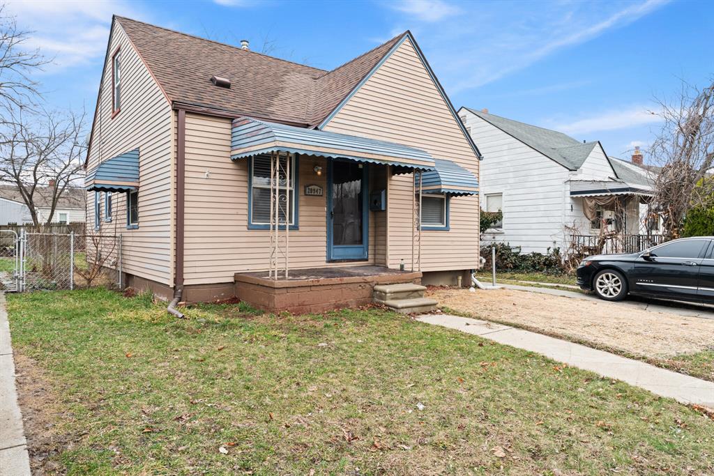 Front exterior of a tan Eastpointe bungalow with blue awnings, steep roof, front lawn, walkway, and side driveway.