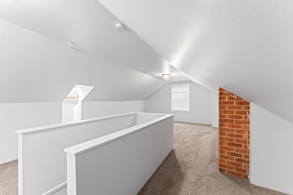 Finished attic with sloped white ceilings, neutral carpet, central stair enclosure, end window, and exposed brick chimney.