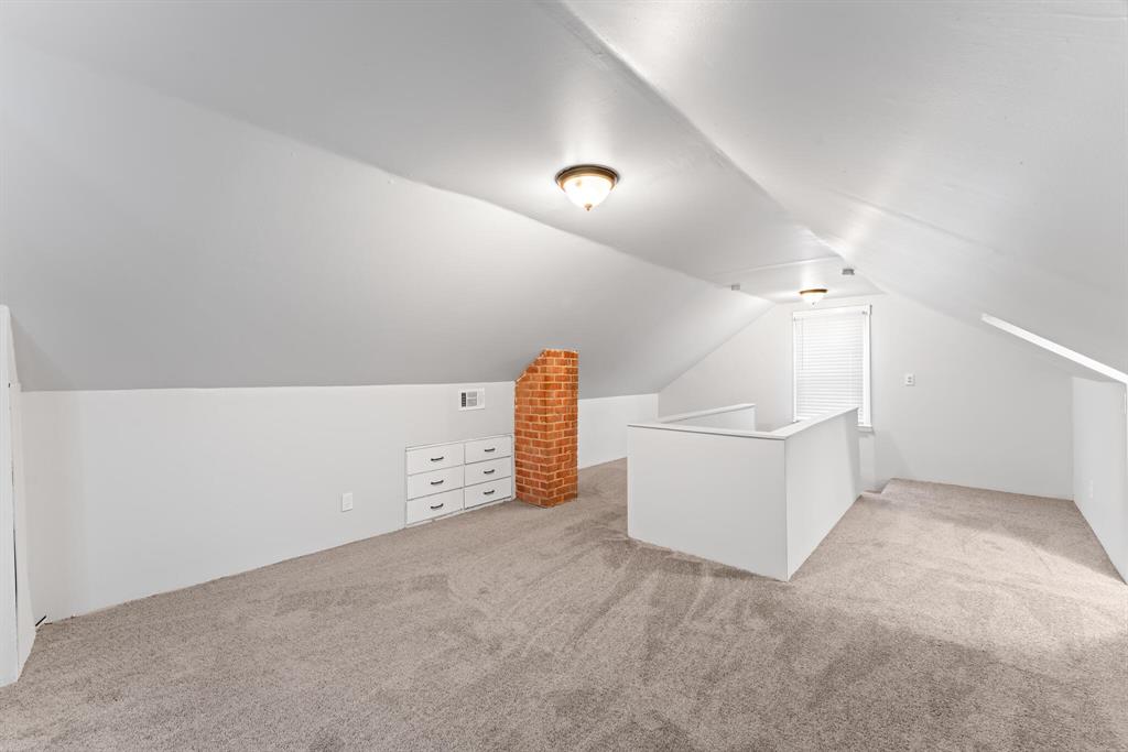 Long finished attic with built-in drawers, sloped ceilings, neutral carpet, stair opening, window, and brick chimney.