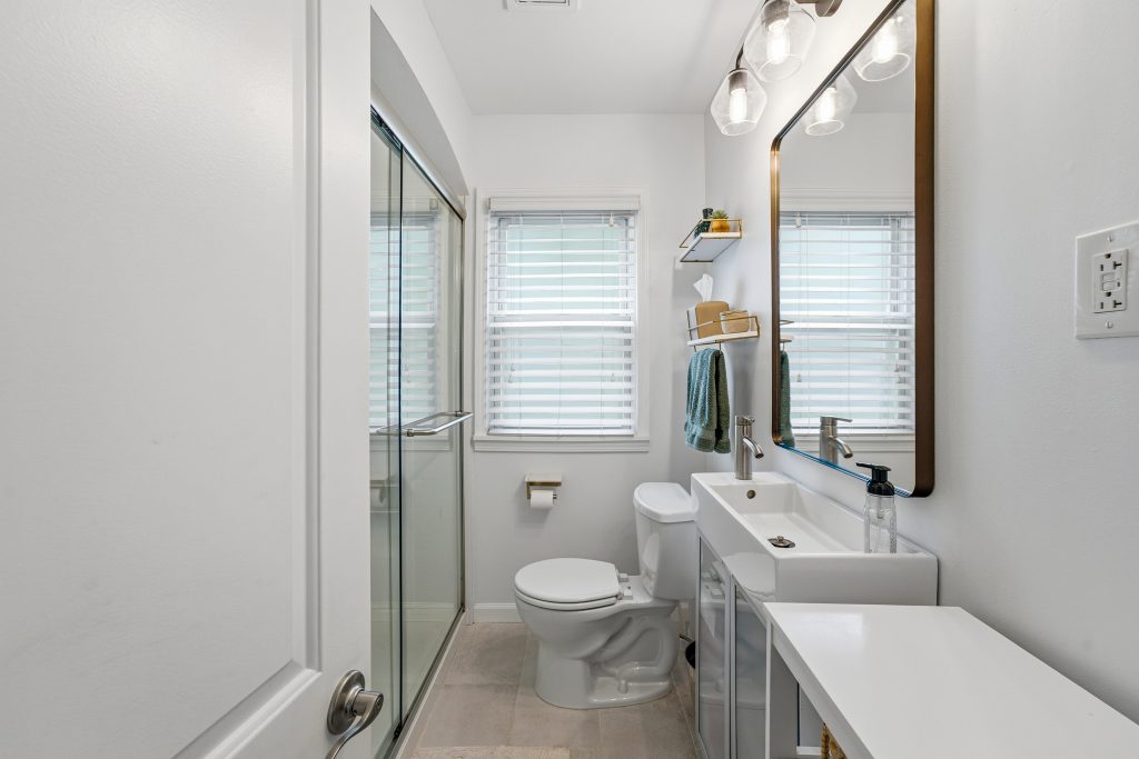 Main-floor bathroom in a Royal Oak home with glass shower, white vanity, large mirror, and window.
