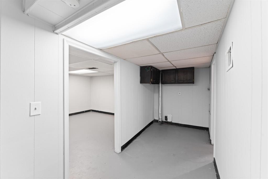 Basement passage with gray painted floor, paneled walls, drop ceiling, fluorescent lighting, storage cabinet, and exposed duct.