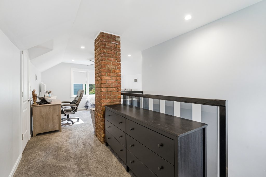 Upper-level bedroom in a Royal Oak home with sloped ceilings, exposed brick chimney, desk area, railing, and carpet.