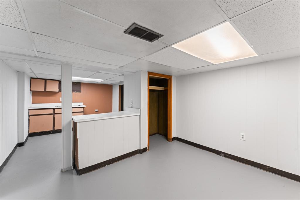 Partially finished basement room with built-in counter, dark cabinets, open closet, support post, gray floor, and drop ceiling.