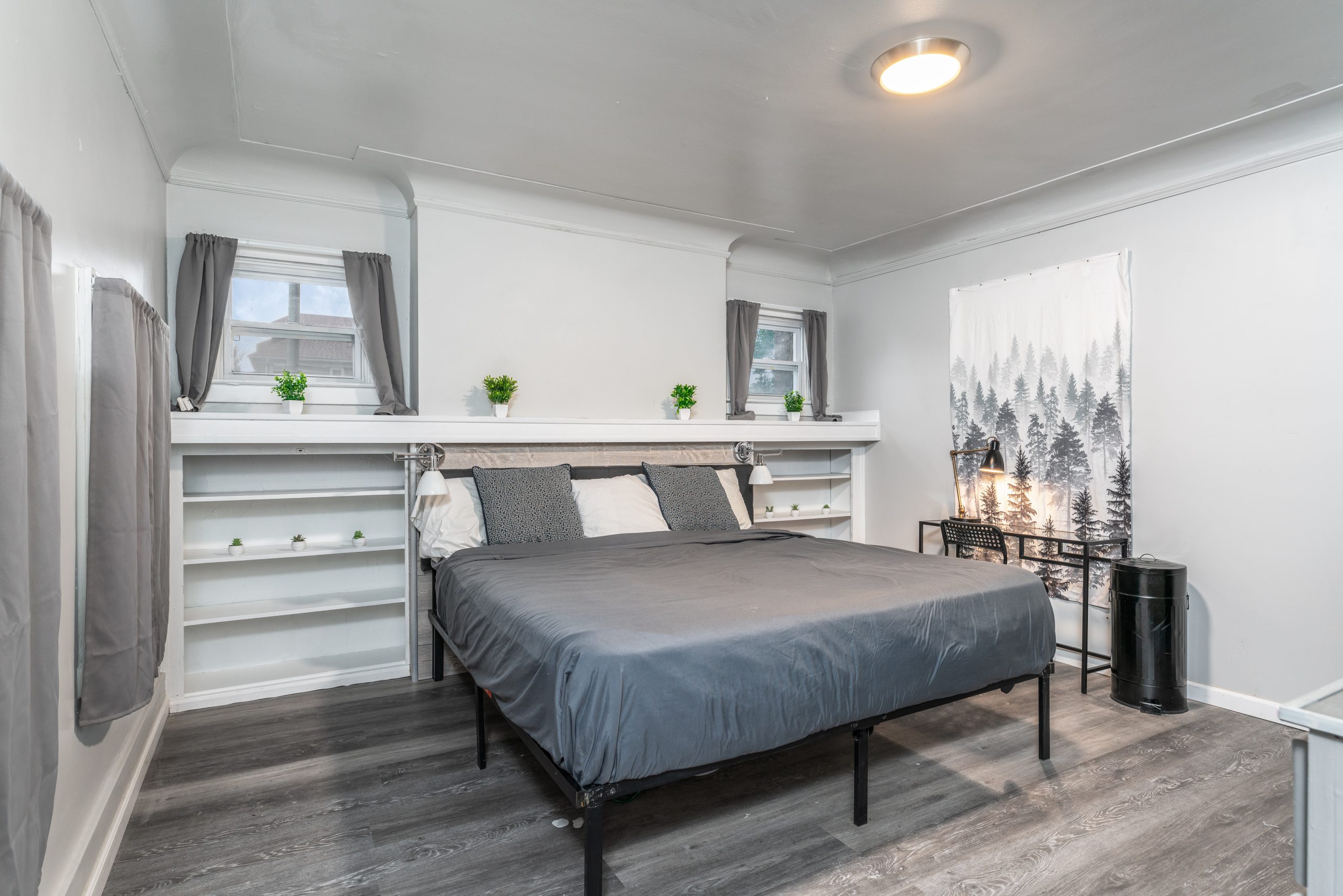 Detroit short-term rental bedroom with gray bedding, built-in wall shelving, two windows, desk, and wood-look flooring.