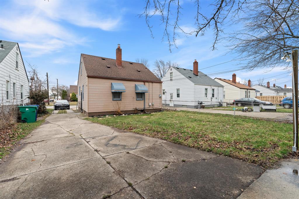 Rear exterior of tan Eastpointe bungalow with blue awnings, brick chimney, concrete driveway, fenced lawn, and neighboring homes.