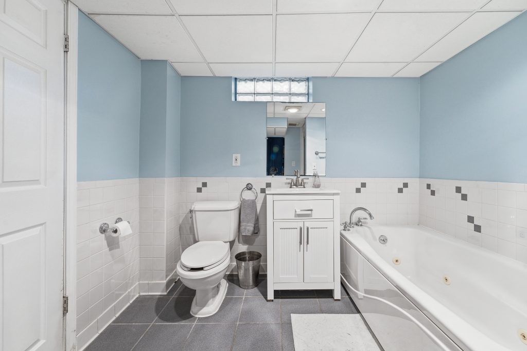 Royal Oak basement bathroom with jetted tub, toilet, white vanity, blue walls, tile, and glass-block window.