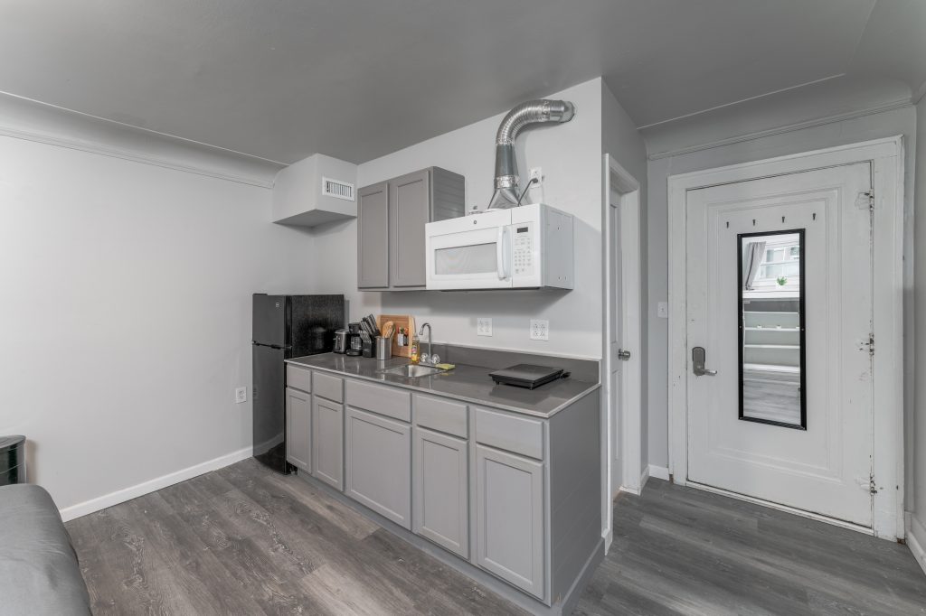 Compact kitchenette in a Detroit guest suite with gray cabinets, sink, microwave, refrigerator, countertop appliances, and private entry door.