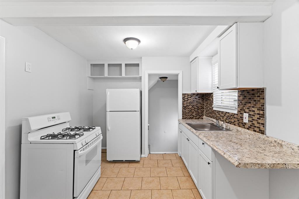 Galley kitchen with white appliances and cabinets, mosaic tile backsplash, stone-patterned counter, tan floor tile, and sink window.