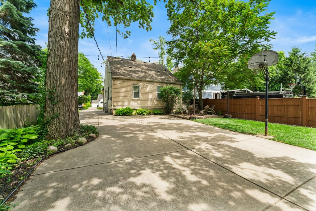 Rear view of a Royal Oak home with long concrete driveway, mature trees, patio, basketball hoop, privacy fence, and rear exterior.
