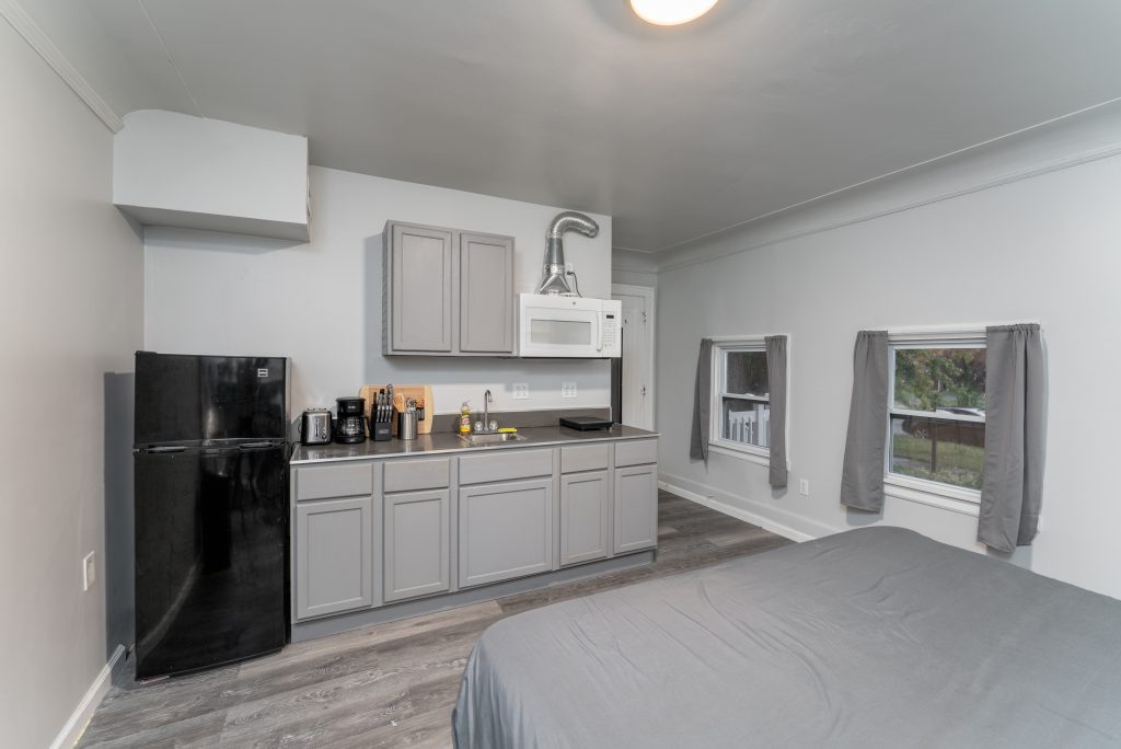 Studio-style Detroit short-term rental showing a bed, gray kitchenette, refrigerator, multiple windows, and wood-look flooring.