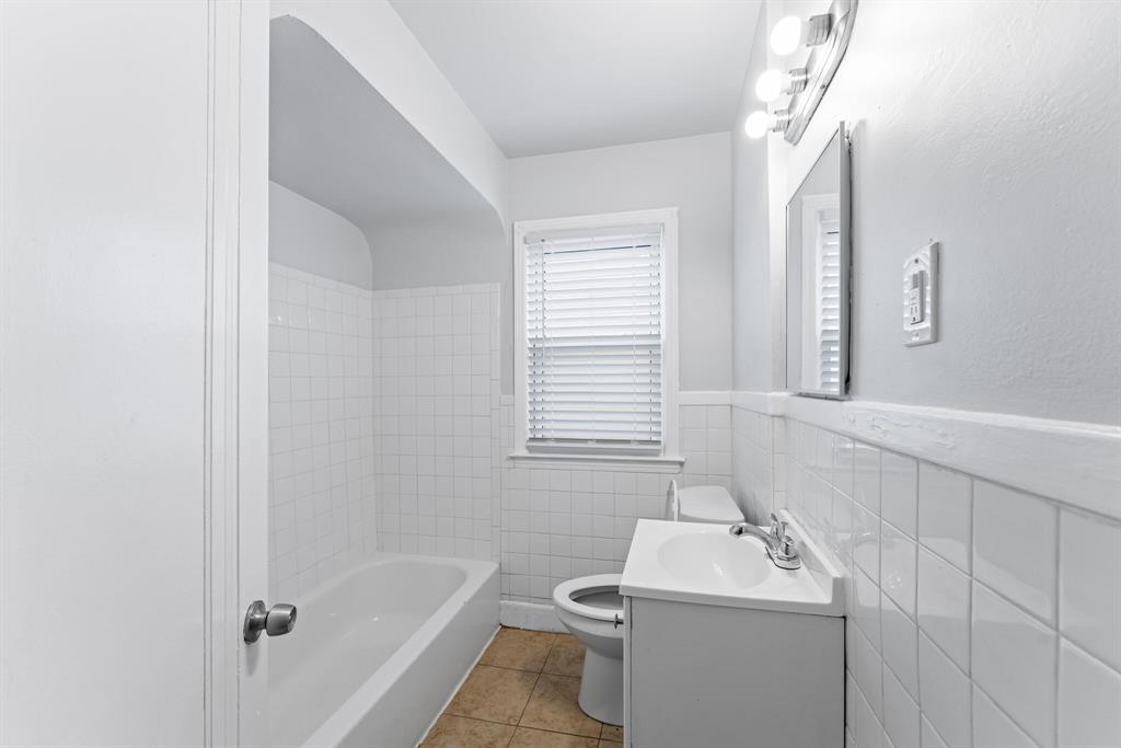 Narrow bathroom with white tub and tile surround, gray walls, tan floor tile, compact vanity, toilet, and window.
