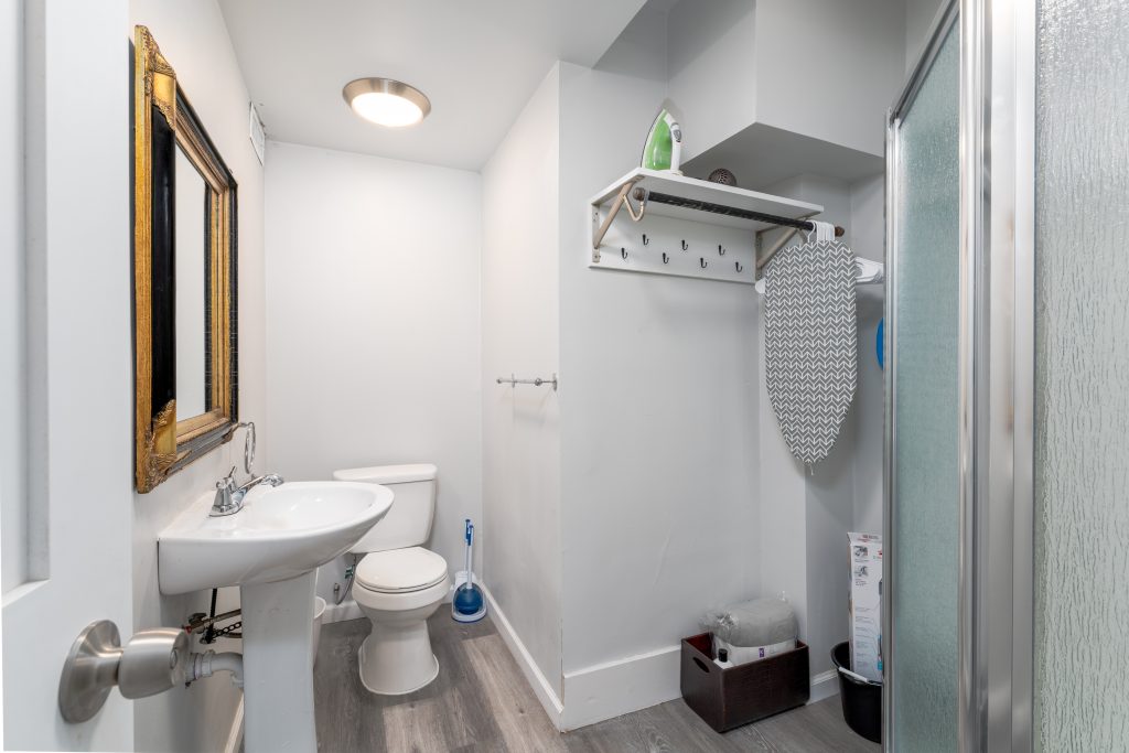 Private bathroom in a Detroit short-term rental with pedestal sink, toilet, glass-enclosed shower, mirror, and ironing storage area.