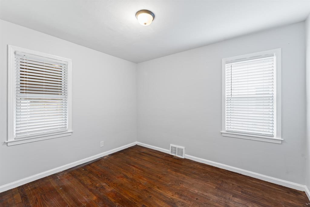 Corner view of an empty bedroom with two windows, hardwood floors, light gray walls, and white trim.