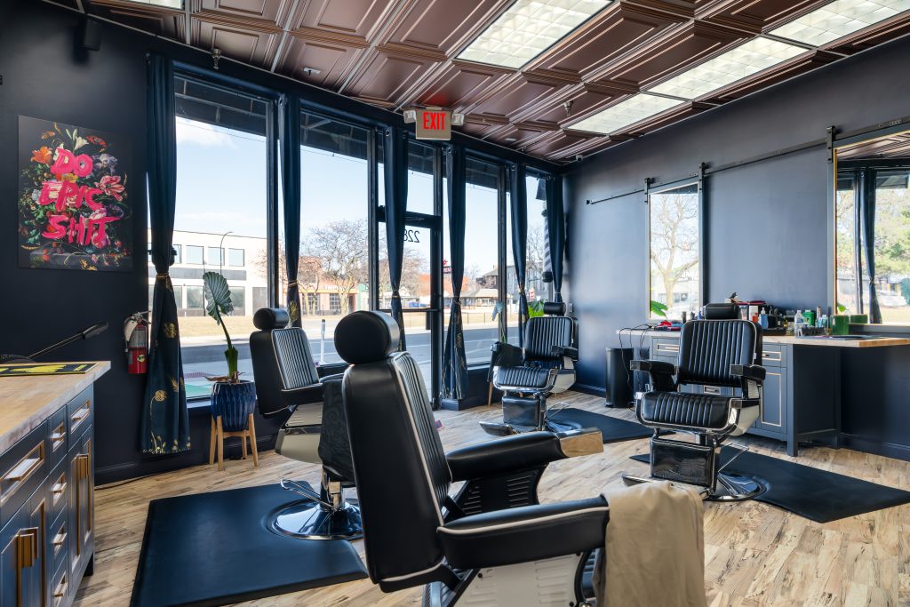 Interior of The Braxton Grooming Lounge in Ferndale with black barber chairs, deep blue walls, brass-accented cabinetry, coffered ceiling, and large storefront windows.