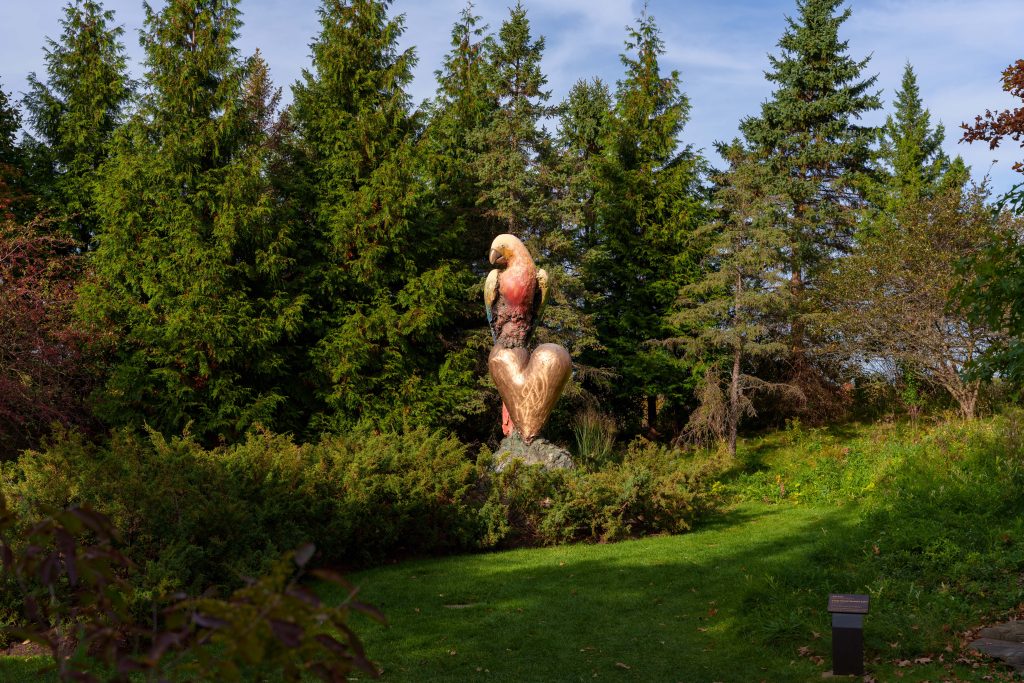 A colorful bronze parrot perches above a golden heart-shaped form, surrounded by tall evergreen trees.
