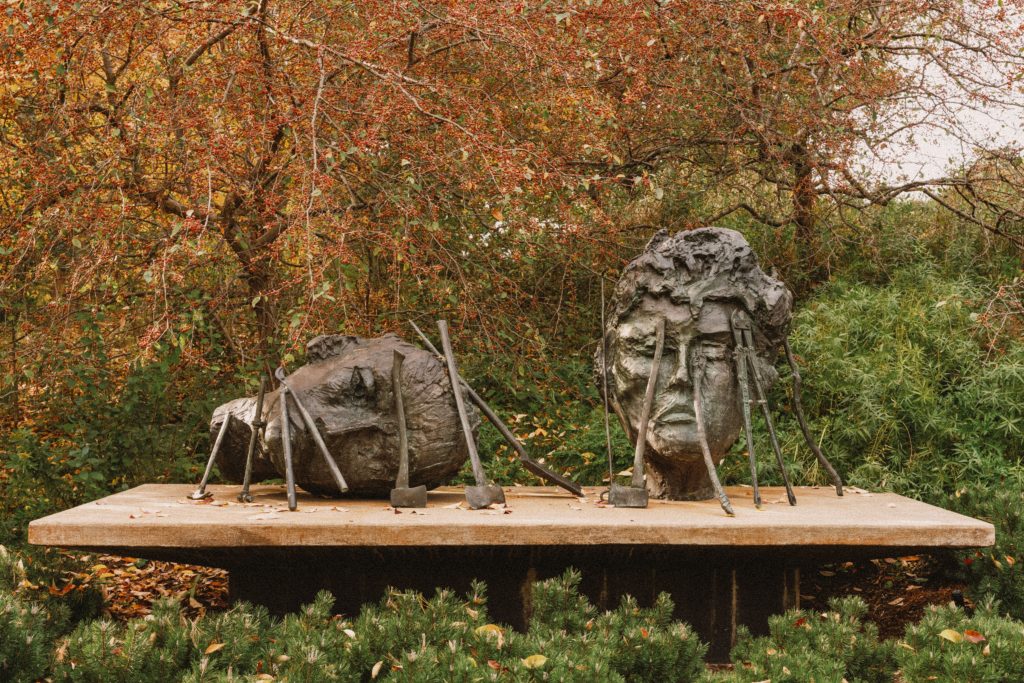 Two roughly textured bronze heads are surrounded by elongated metal tools on a platform beneath orange autumn foliage.