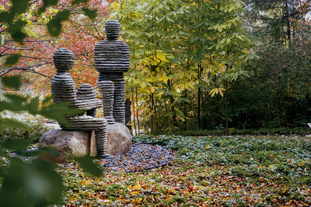 Layered bronze human figures sit against natural boulders, surrounded by fallen leaves and dense woodland vegetation.