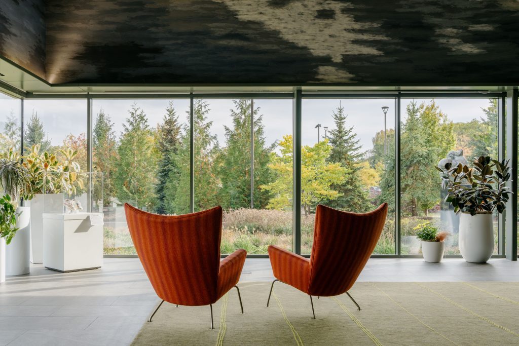 Two rust-colored lounge chairs face floor-to-ceiling windows overlooking autumn trees at Frederik Meijer Gardens.